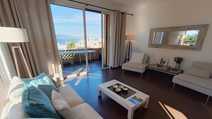 A modern living room with a white sofa, a coffee table, and a balcony with a view of the sea and buildings. - Apartment Grace by Interhome (Saint-Raphaël)