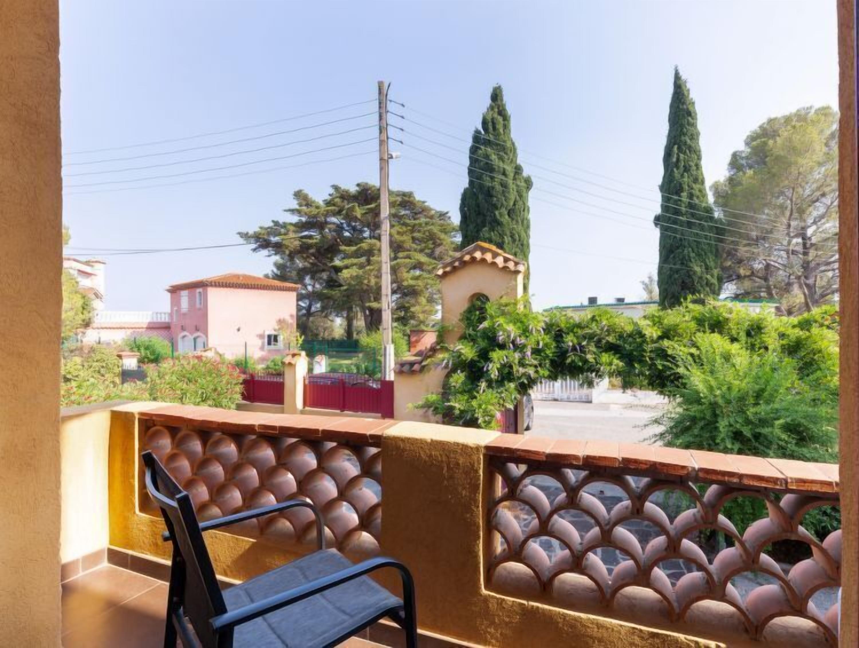 Un balcón con una silla, una barandilla de metal decorativa, y vistas a casas y árboles vecinos.