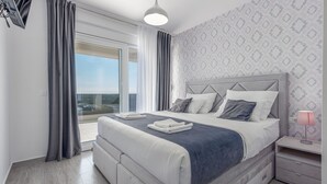 A modern bedroom with a large bed, a nightstand, a lamp, and a view of the outdoors. - Villa Suton (Razanac)