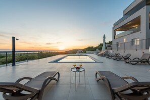 A rooftop pool area with lounge chairs and a table with drinks, overlooking a sunset and a distant view. - Villa Suton (Razanac)