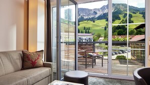 A modern living room with a sofa, a coffee table, and a view of a mountain landscape through large glass doors. - Panoramahotel Oberjoch (Bad Hindelang)