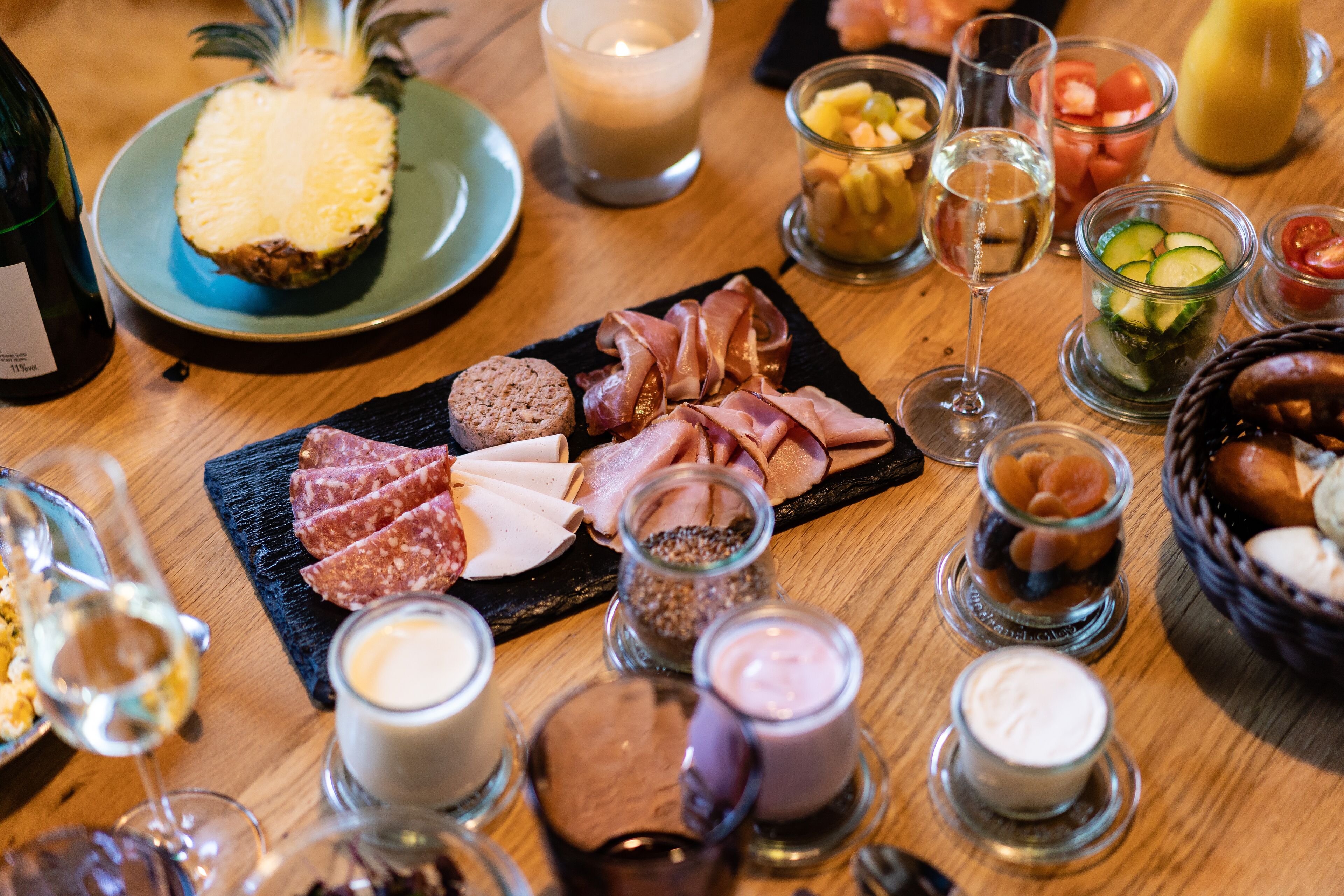 Una mesa de madera con diversos alimentos, incluyendo carnes, quesos y frutas, junto con varios vasos de bebidas.