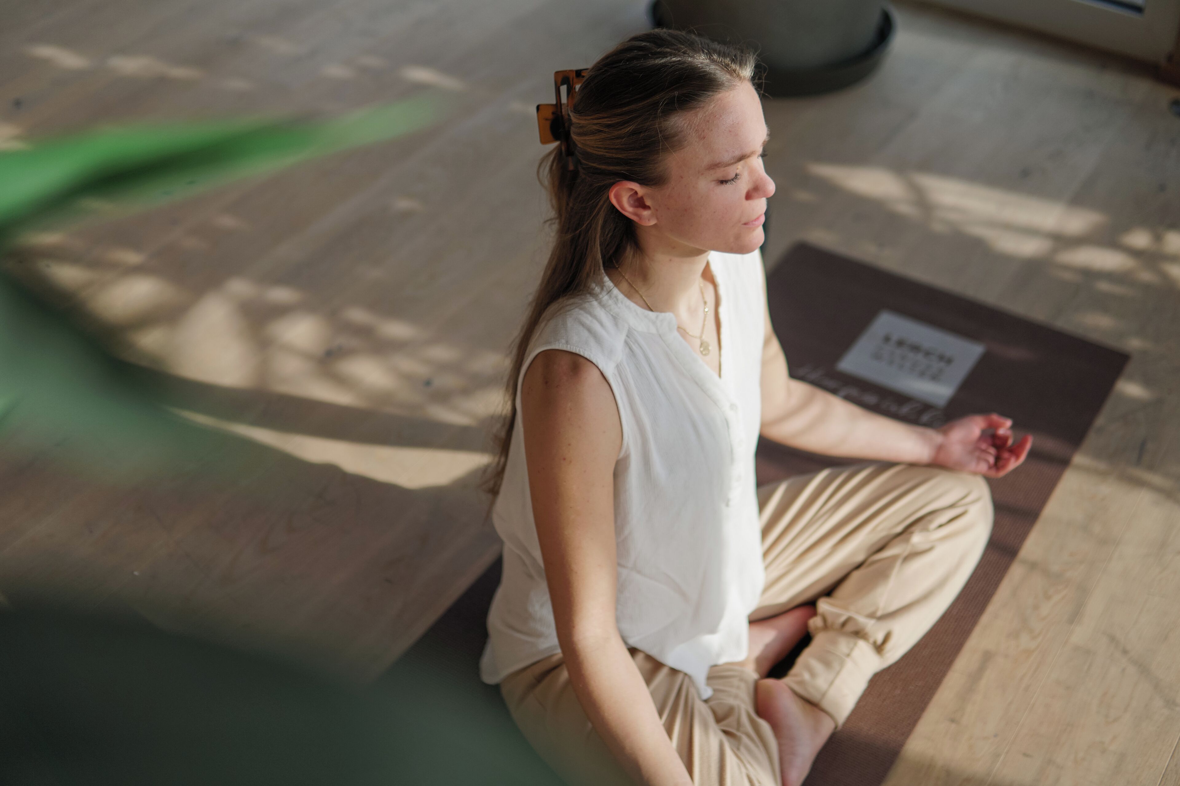 A person sitting on a mat in a room with wooden flooring and a visible yoga mat.