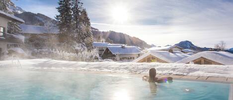 Una piscina cristalina con una persona nadando, rodeada de edificios y árboles cubiertos de nieve bajo un cielo soleado.