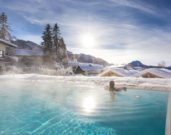 A clear pool with a person swimming, surrounded by snow-covered buildings and trees under a sunny sky.