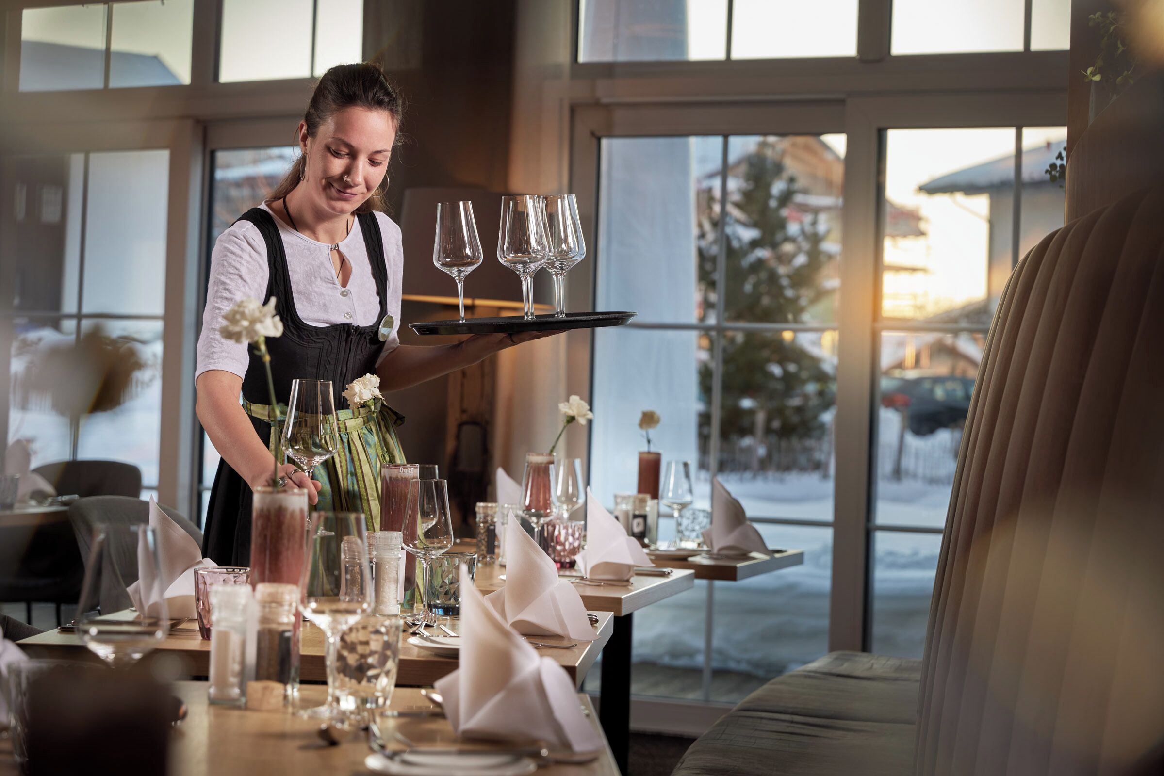 Une scène de restaurant : un serveur tient un plateau rempli de verres, une table est dressée avec différents plats, et de grandes fenêtres offrent une vue sur un paysage enneigé.