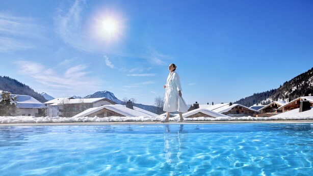 A person in a white robe walking by a clear blue pool with snow-covered buildings in the background. - Panoramahotel Oberjoch (Bad Hindelang)