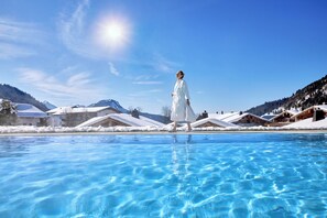 Eine Person in einem weißen Bademantel geht an einem klaren, blauen Pool entlang, im Hintergrund befinden sich schneebedeckte Gebäude.