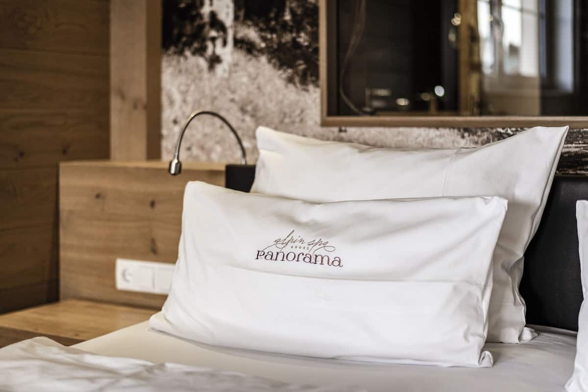 Een netjes opgemaakt bed met witte lakens en een kussen met de tekst 'Alpin Style Panorama'.