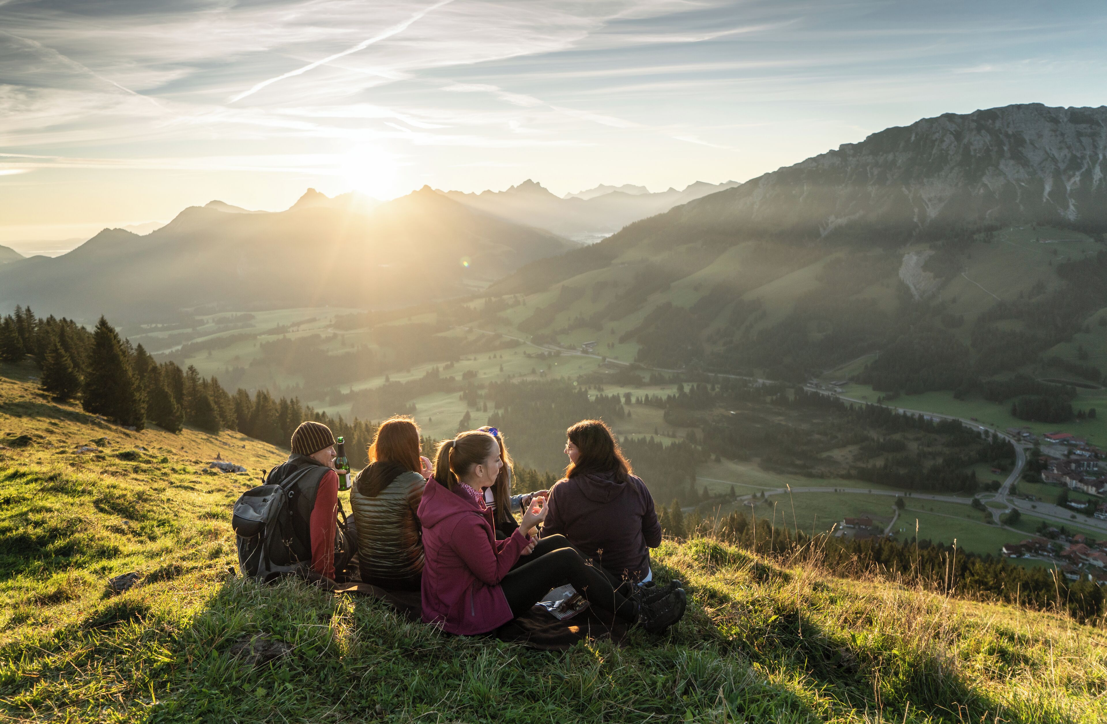 A group of people sitting on a grassy hill overlooking a valley with mountains in the background.