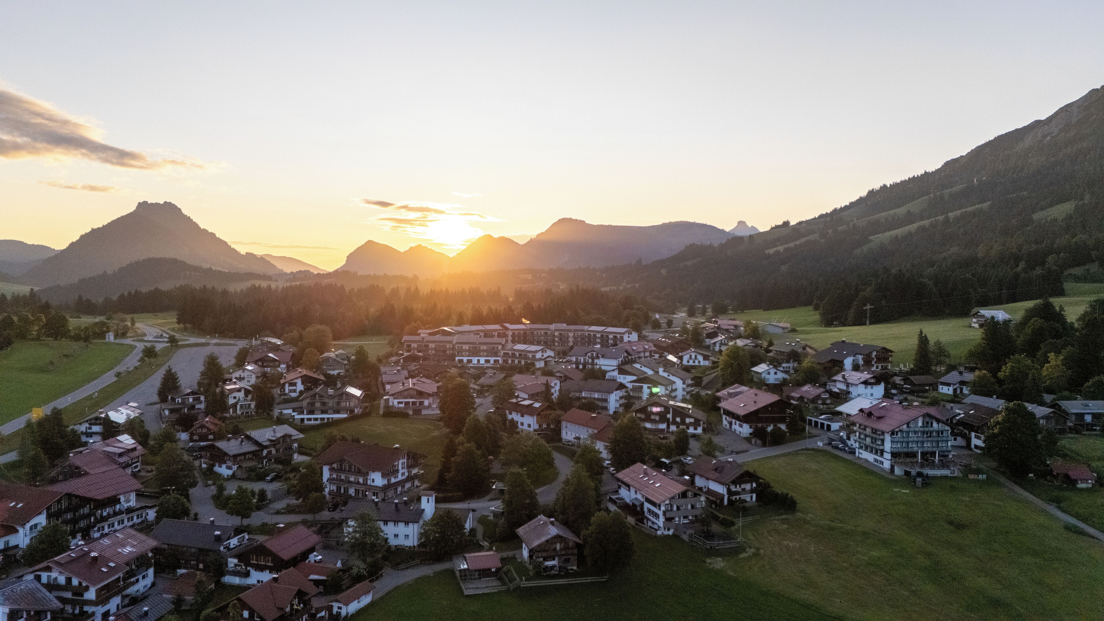 A residential area with houses and a road, surrounded by mountains and a sunset.