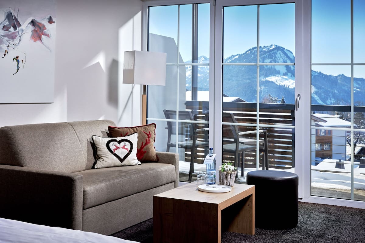 Een moderne hotelkamer met een bank, een klein tafeltje en uitzicht op besneeuwde bergen door grote ramen.