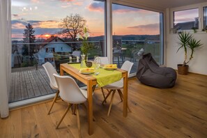 A dining area with a table set for two, a bean bag chair, and a view of the sunset through large windows.