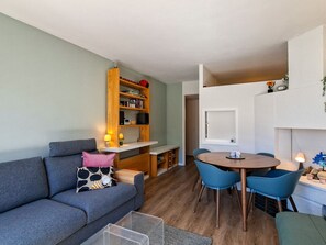 Un espacio habitable compacto con sofá, una mesa de comedor con sillas, una estantería y una pequeña zona de cocina.