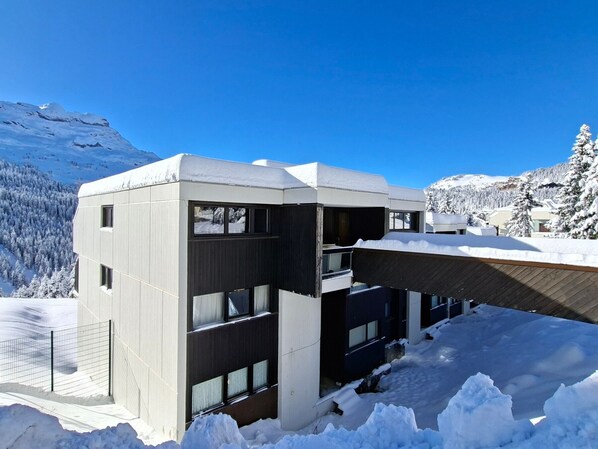A modern building with a flat roof, large windows, and a snow-covered landscape in the background.
