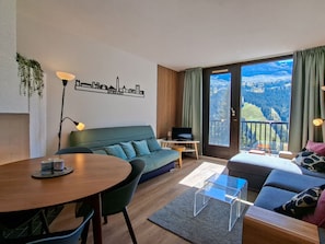 A modern living room with a sofa, a dining table, and a TV. There is a balcony with a view of the outdoors.