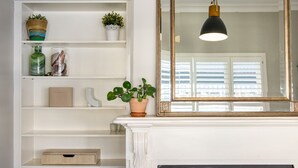 Un interior blanco con chimenea, un espejo grande con marco dorado y estanterías con diversos objetos, incluyendo plantas y adornos.