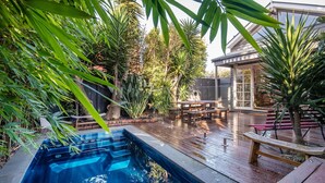 Patio trasero con piscina, terraza de madera y zona de estar exterior rodeada de abundante vegetación.