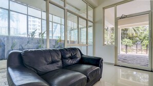 A modern living room with a black leather sofa, large windows, and a view of greenery outside. - Itsy Hotels Ushodaya Royal Residency (Thiruvananthapuram)