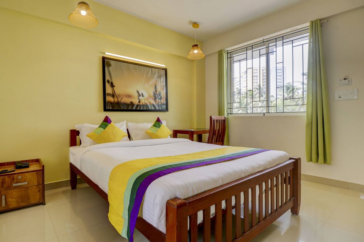 Habitación de hotel con cama, mesitas de noche, un cuadro, ventana con cortinas y vistas a edificios.