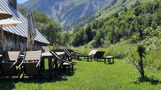 A rustic outdoor seating area with wooden tables and chairs, a stone chimney, and a view of green mountains. - Comfort 2-room, 6 ppl, Terrace & Garden in Valloire (Valloire)