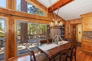 A wooden interior with a dining table, chairs, and a chandelier. There is a large window with a view of trees and a deck outside.