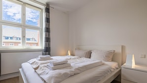 Un lit bien fait, avec du linge de lit et des serviettes blanches, une table de chevet avec une lampe et une fenêtre donnant sur des immeubles.