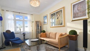 A living room with a blue chair, beige sofa, glass coffee table, and artwork on the walls.