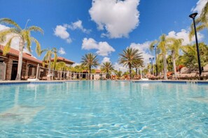 A clear blue pool surrounded by palm trees and lounge chairs, with a building featuring a red roof in the background. - The Cinehaven at Solterra by Shine Villas 407 (Davenport)
