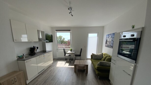 A modern kitchen with white cabinets, a dining area with a green sofa, and a large window providing a view of the city. - City Apartments Wilhelmshaven (Wilhelmshaven)