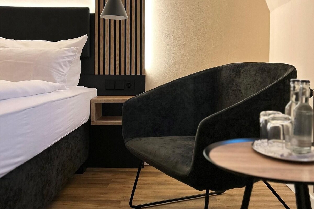 Een moderne hotelkamer met een bed, een donkere fauteuil, een klein tafeltje met glazen en een wandlamp.