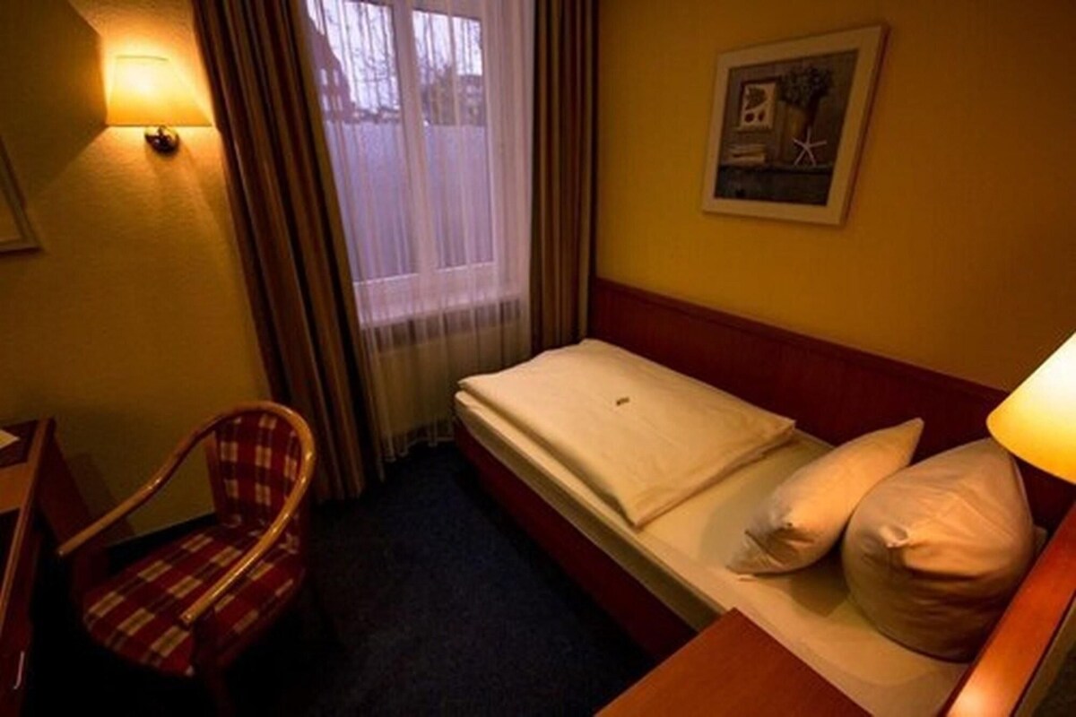 Een hotelkamer met een enkel bed, een stoel, een raam met gordijnen en een schilderij aan de muur.