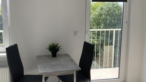 A small balcony with a table and two chairs, a potted plant, and a view of greenery outside. - City Apartments Wilhelmshaven (Wilhelmshaven)