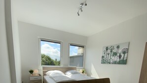 A bedroom with a bed, a sofa, a nightstand, a lamp, and a window. - City Apartments Wilhelmshaven (Wilhelmshaven)