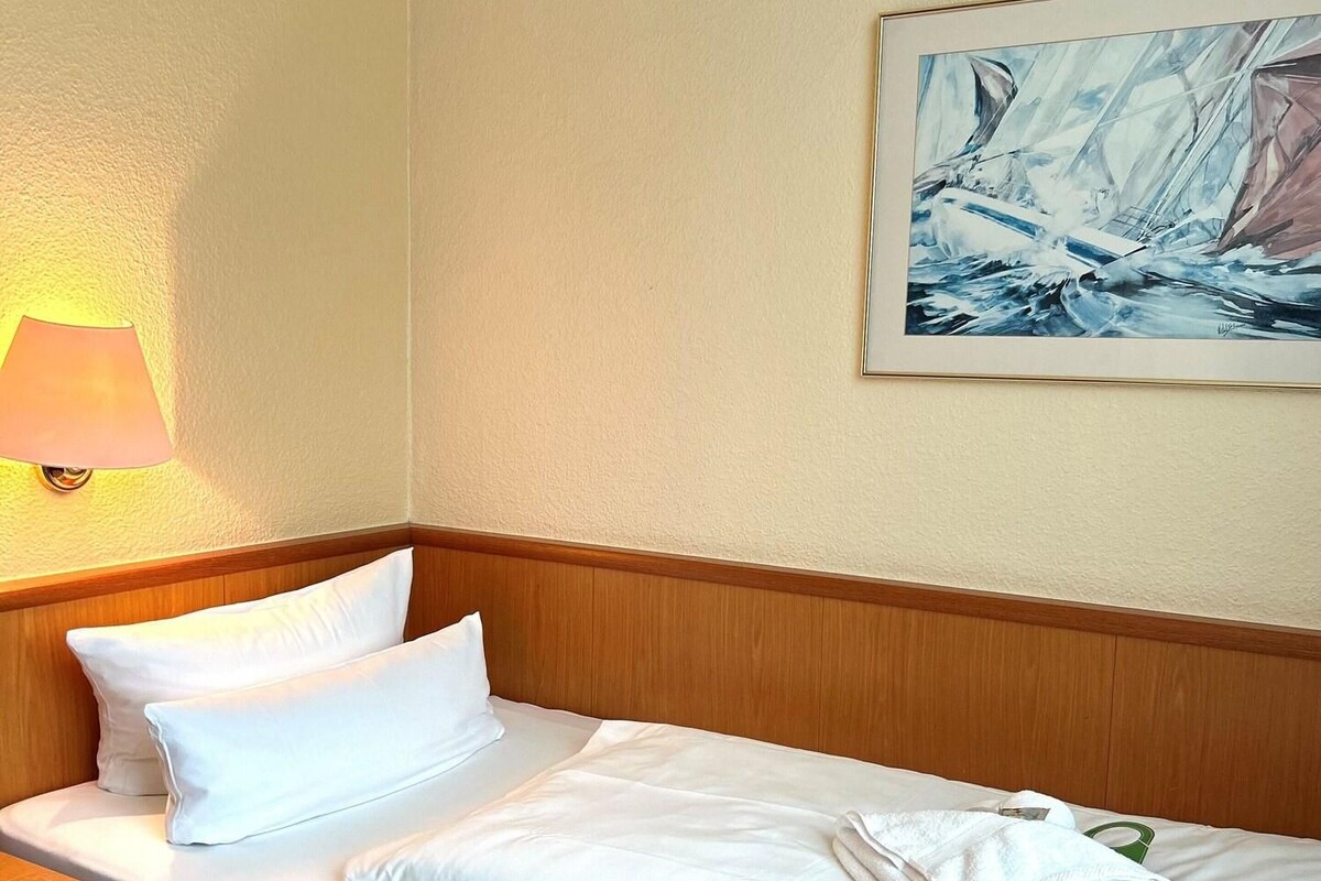 Een keurig opgemaakt bed met witte lakens en kussens, een houten hoofdbord, een schilderij van een zeilboot aan de muur en een bedlampje.