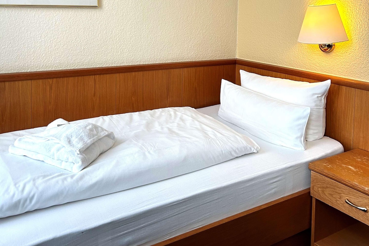 A neatly made bed with white linens and pillows, a wooden headboard, a bedside table with a lamp, and a wooden nightstand.