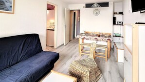 A compact living space with a dark blue sofa, a woven pouf, a dining table with chairs, a kitchen area with a TV, and a clock on the wall.