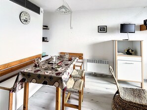 A dining area with a table set for four, a clock on the wall, a television, and a cabinet with a picture frame. - Functional studio 28m² · Ski-in · Pool access (Le Corbier)