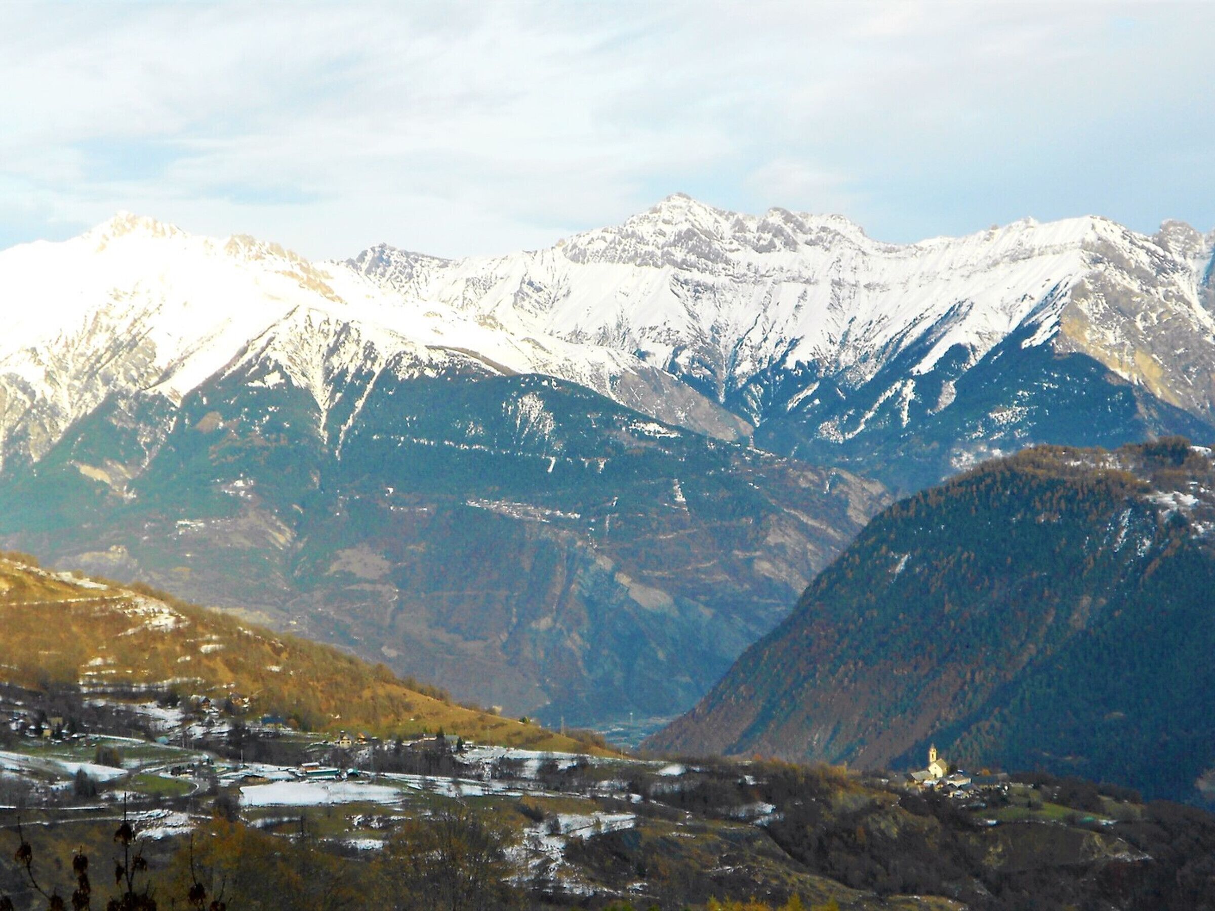 Eine Berglandschaft mit schneebedeckten Gipfeln und einem Tal mit einer kleinen Siedlung.