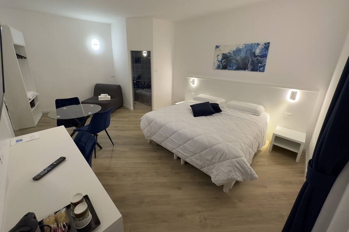 A modern hotel room with a bed, bedside tables, a desk with a chair, a small table, and a mirror.