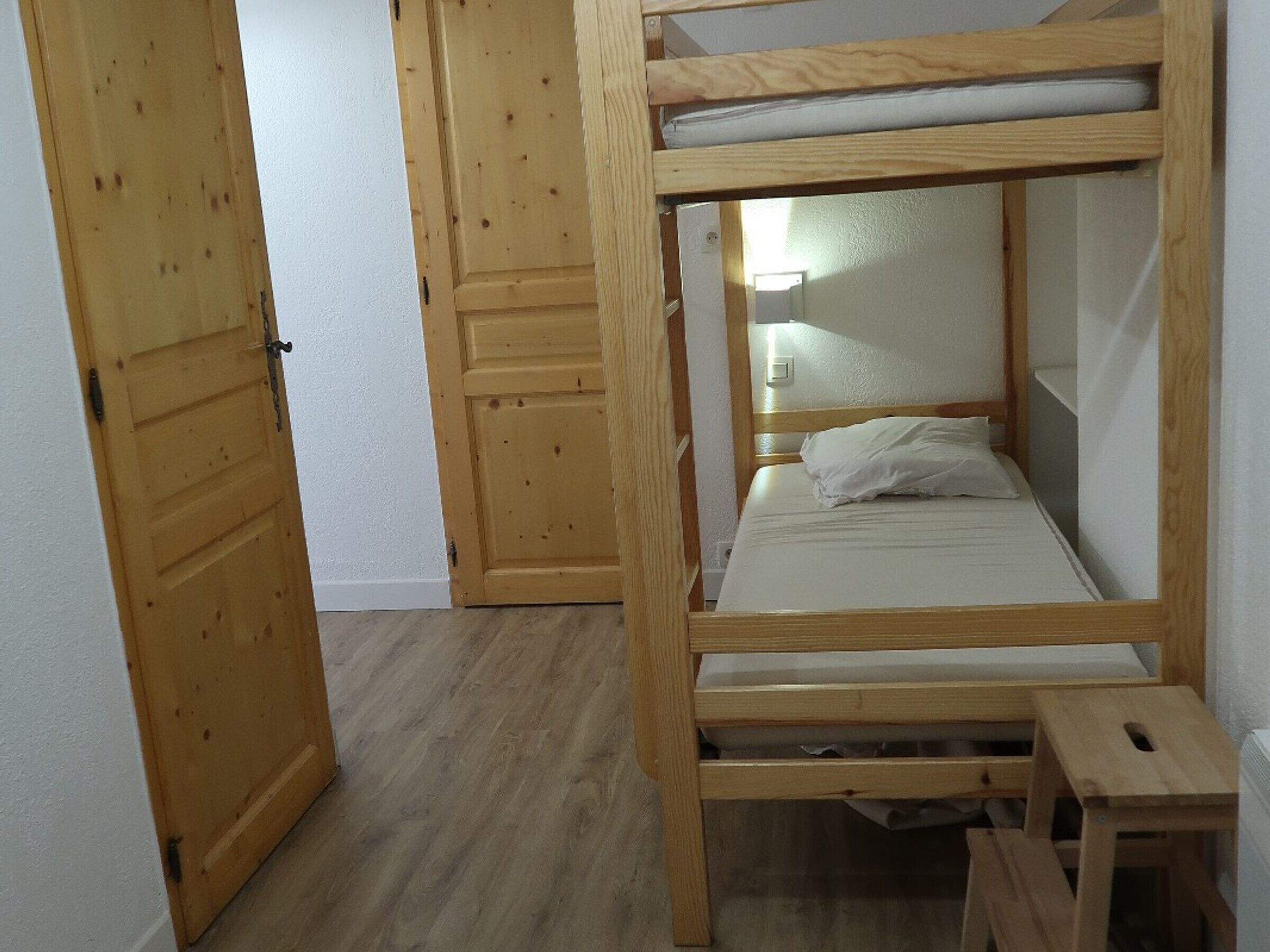 A bunk bed with a wooden frame, a small wooden stool, and a white mattress.