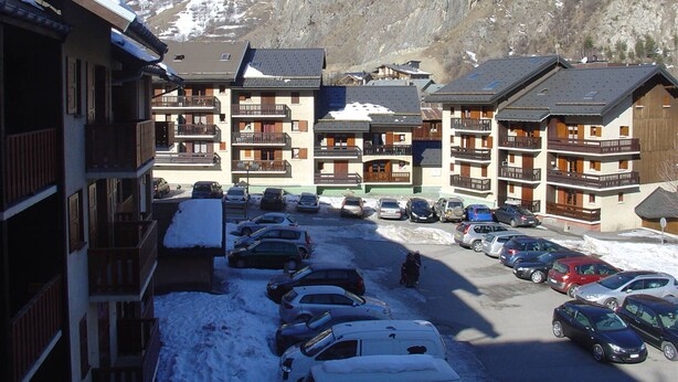 A mountain resort with multi-story buildings, a parking lot, and snow-covered mountains in the background. - 2-room cabin apartment for 4 people, west facing with balcony and collective parking (Valloire)