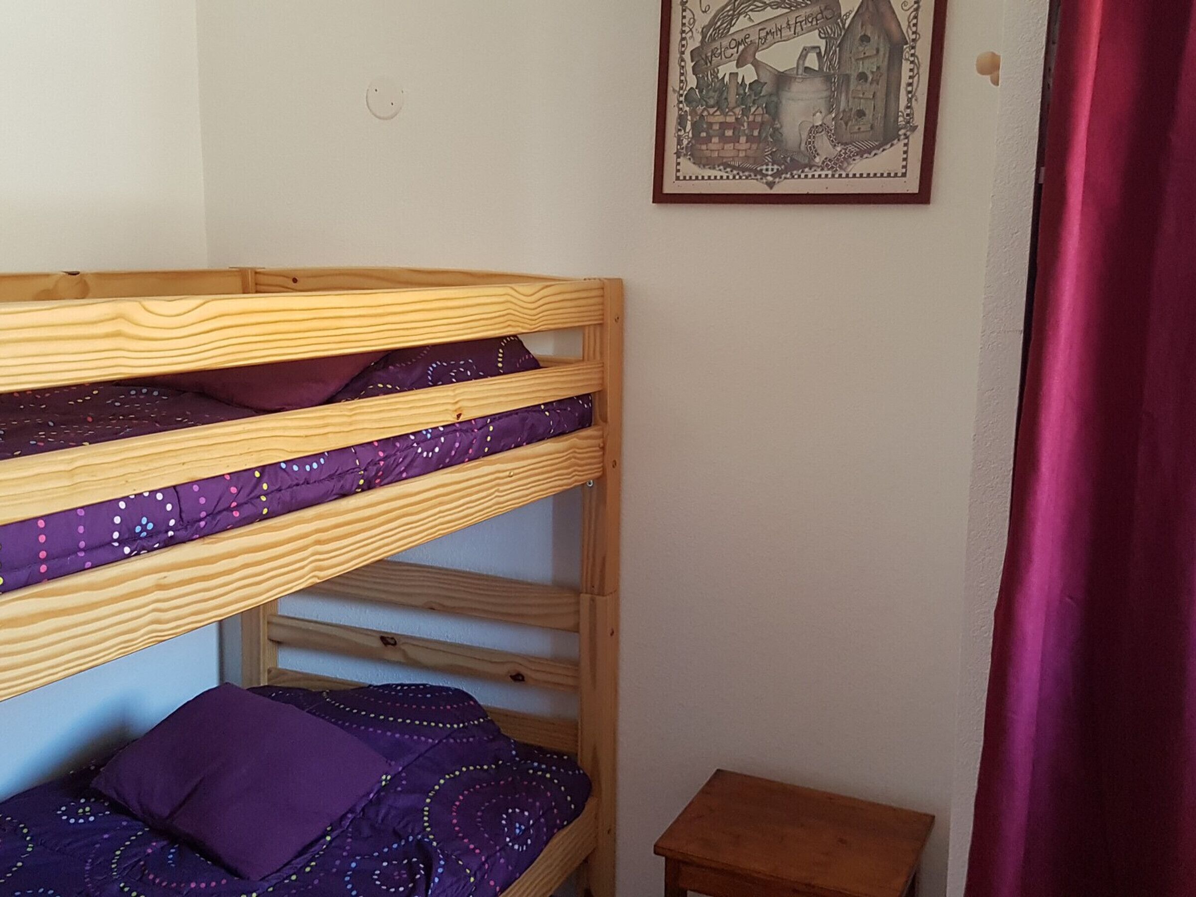 Una cama litera con ropa de cama púrpura, una pequeña mesa de madera y un cartel de bienvenida en la pared.