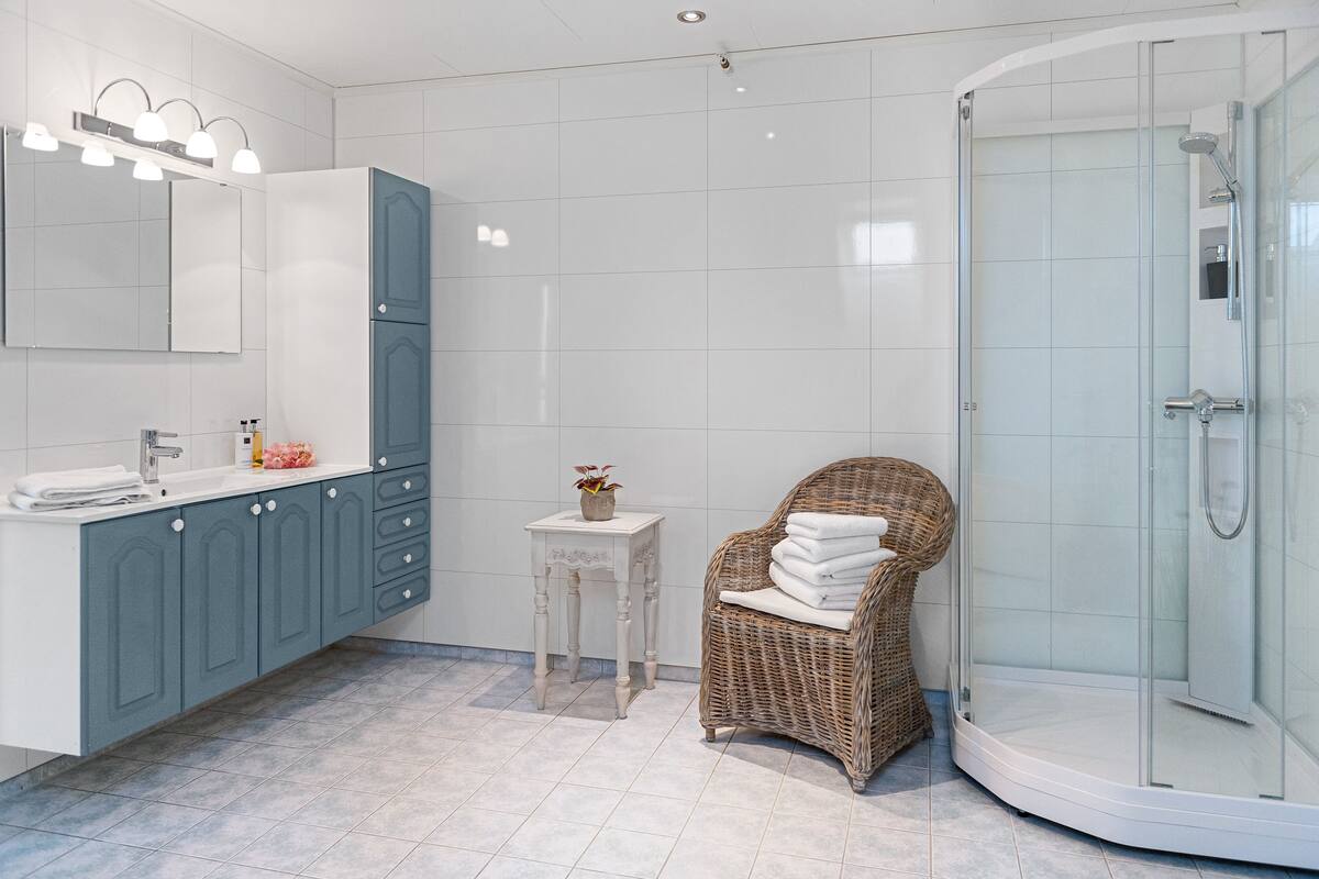 A modern bathroom with a glass-enclosed shower, a wicker chair with towels, and a vanity with a mirror and lights.