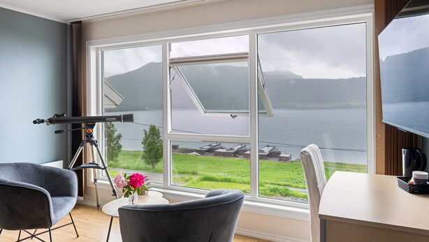 A room with a large window overlooking a body of water and mountains, a telescope, and modern furniture. - Moens Motell (Vanylven)