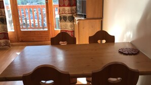 A wooden dining table with four chairs, a television on a wooden cabinet, and a view of a wooden deck through a window.
