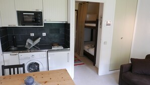 Une cuisine compacte équipée d’un four à micro-ondes, d’un four et d’un lave-linge. Une petite table à manger, agrémentée d’un objet décoratif, se trouve au premier plan. On aperçoit un lit superposé dans la pièce adjacente.