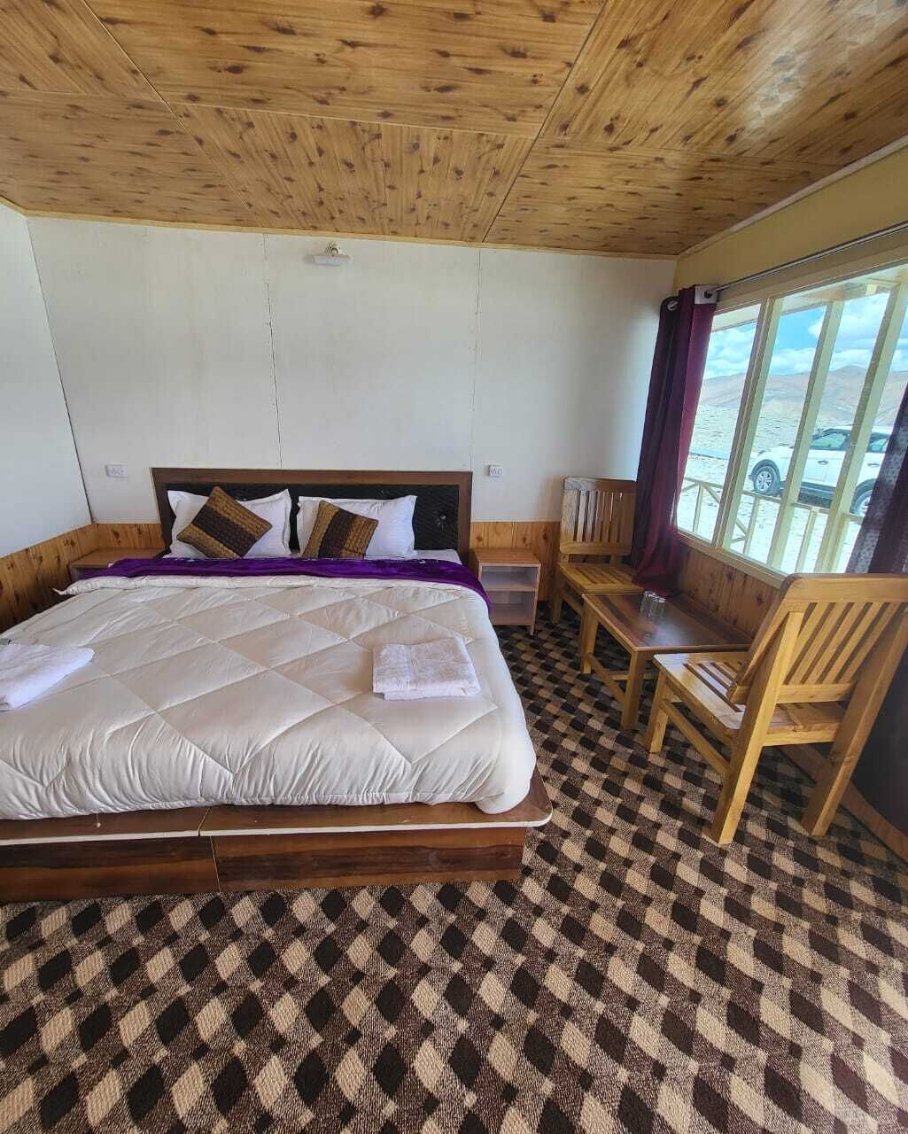 Una habitación con una cama, cabecera de madera, suelo de cuadros, ventana con vista al exterior y una silla de madera.