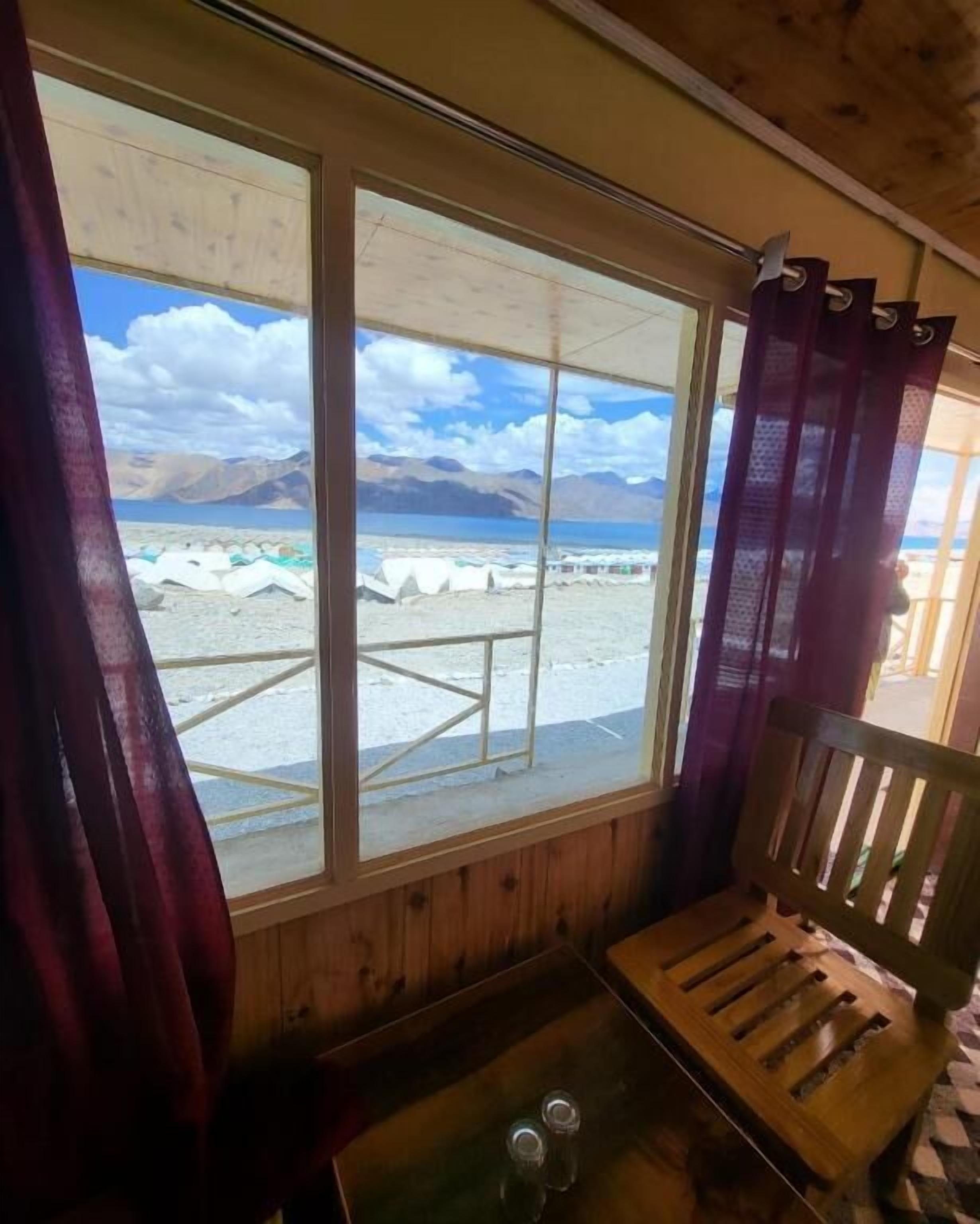 Een houten vakantiehuis met een groot raam met uitzicht op een strand en bergen.