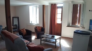 A living room with a sofa, two armchairs, a coffee table, and a TV. There are windows with curtains, a radiator, and a trash bin.